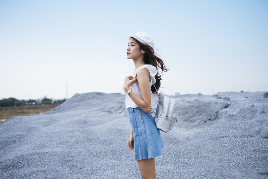 美丽的亚泰旅行家妇女戴着帽子和白色袋子站在岩石山的侧视图。刮风的日子。