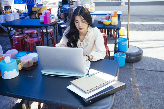 穿着休闲衬衫的泰国工作女商人在街头小吃摊用笔记本电脑在户外工作。