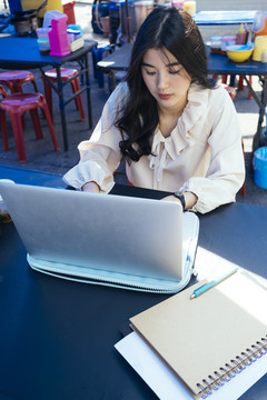 穿着休闲衬衫的泰国工作女商人在街头小吃摊用笔记本电脑在户外工作。