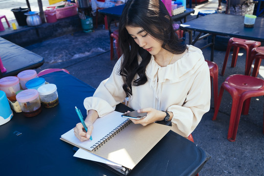 穿着休闲衬衫的美丽泰国职业女商人记下顾客的订单。使用智能手机查看电子邮件并与人联系。