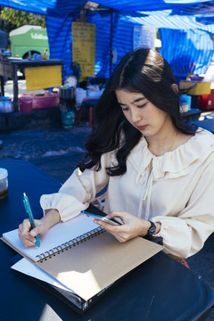 穿着休闲衬衫的美丽泰国职业女商人记下顾客的订单。使用智能手机查看电子邮件并与人联系。
