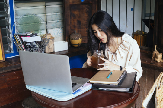 穿着休闲衬衫在家工作的泰国美女。用玻璃杯喝可乐和苏打水。