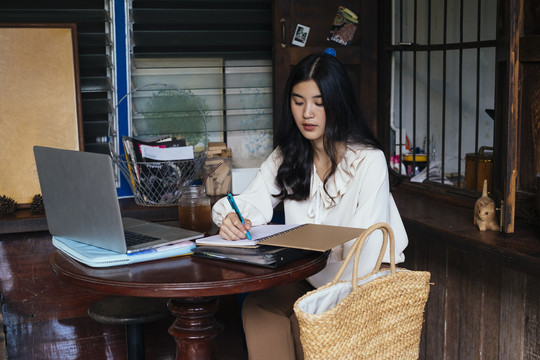 穿着休闲衬衫的泰国工作女商人正在笔记本电脑前用笔记笔记。