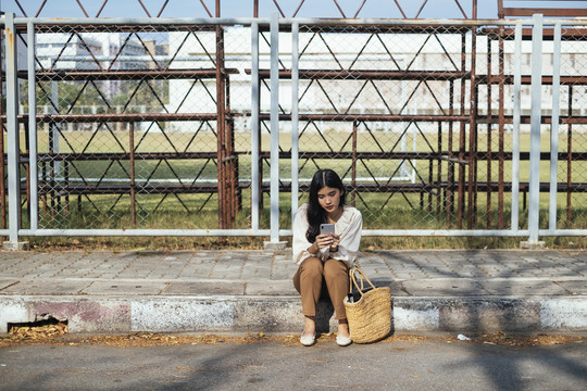 坐在人行道上使用智能手机的泰国年轻女子。