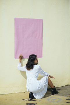 黑色卷曲的长发女孩身着白色连衣裙，以别致的姿态坐在黄色墙面上，用粉色的纸，复制空间。