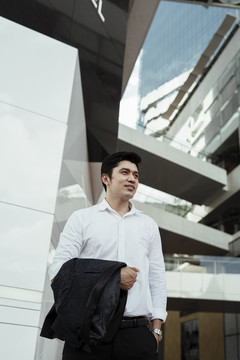 一个成熟的亚洲商人，穿着白色正式衬衫，胳膊上挽着西装，站在城外。