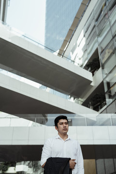 一个成熟的亚洲商人，穿着白色正式衬衫，胳膊上挽着西装，站在城外。
