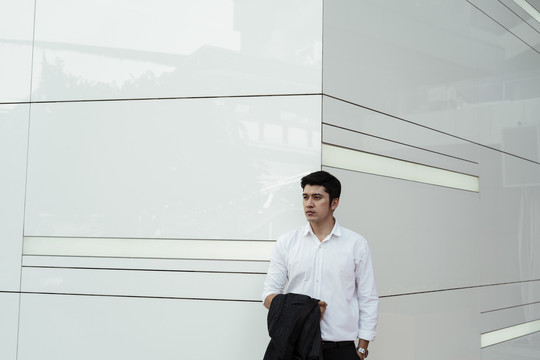成熟的亚洲商人穿着白色正式衬衫，胳膊上挽着西装，站在大楼角落的白墙上。