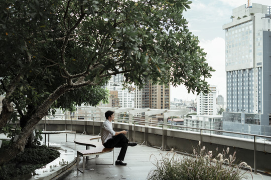一个穿着白色正式衬衫的成熟的亚洲商人坐在屋顶的长凳上，在大树下看着城市。