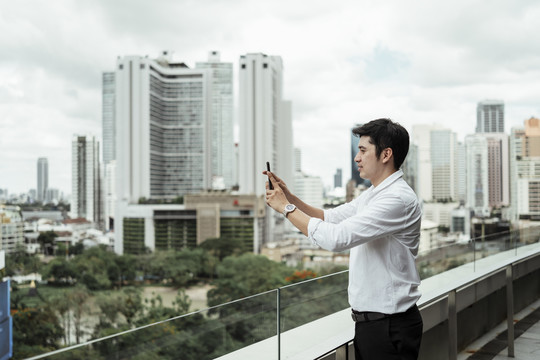 穿着白色正式衬衫的成熟亚洲商务人士在屋顶用智能手机拍摄城市景观。