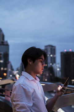 一个穿着白衬衫、戴着眼镜的黑发男人站在屋顶上，看着他的智能手机，以城市夜景为背景阅读朋友的留言。