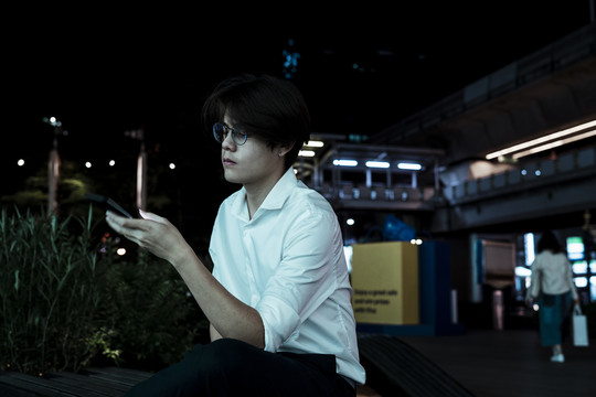黑发男坐在火车站旁边的人行道上，用智能手机。