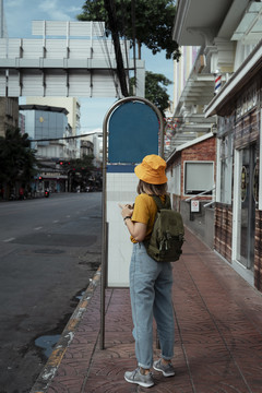 一个穿着黄色衣服和牛仔裤的背包妇女站在城市的路边人行道上，看着公共汽车站上的细节。