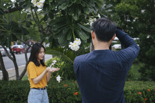 男摄影师拿着鲜花和相机，在鸡蛋花树旁为十几岁的模特女孩拍照。