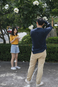 男摄影师拿着鲜花和相机，在鸡蛋花树旁为十几岁的模特女孩拍照。