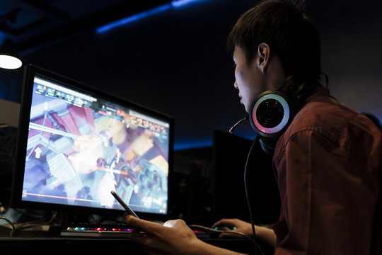 亚裔玩家在网吧玩电脑游戏，戴着耳机，看着显示器屏幕。