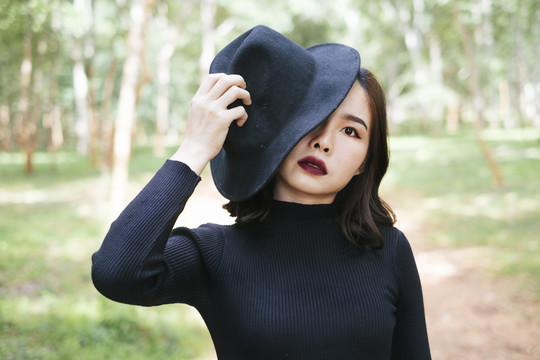 穿黑衣服的女人戴着黑帽子遮住半张脸，站在森林里。