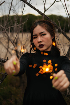一个穿着黑衣服的女人双手拿着两个火花，在枯树中间。