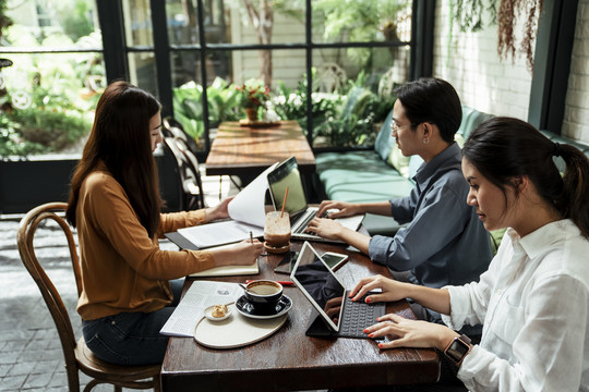 三个年轻的商务人士坐在桌子旁，使用笔记本电脑、平板电脑和电脑。严肃的商务会议。在咖啡馆户外工作。