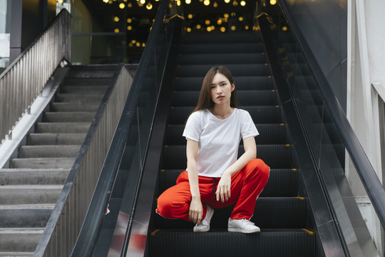 时尚写真——坐在自动扶梯上的穿着白衬衫和红色运动裤的美丽亚洲年轻女子。后面有漂亮的波基背景。