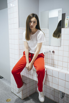 时尚写真-美丽的亚洲年轻女子穿着白衬衫和红色运动裤坐在公共卫生间内的水槽。镜子上的倒影。