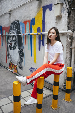 时尚写真-年轻的泰国亚裔女子穿着白衬衫和红色运动裤坐在黄色的障碍在街头小巷。
