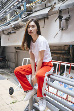 全长时尚肖像-美丽的亚洲年轻女子在白衬衫和红色运动裤坐在铁栏在街头小巷。