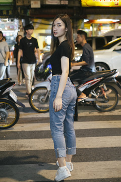 时尚写真-美丽的时髦亚洲年轻女子在黑色衬衫和牛仔裤裤子走在马路对面的夜晚在曼谷。