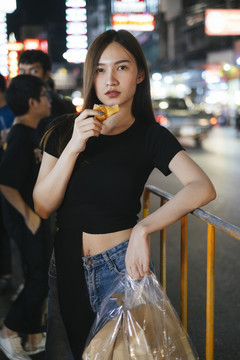 时尚写真-穿着黑色衬衫和牛仔裤裤子的亚洲时髦美女喜欢在曼谷买吃街头食物。夜生活。