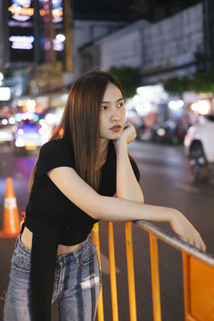 时尚写真——一位穿着黑色衬衫和牛仔裤的亚洲时髦年轻女子斜靠在人行道扶手上。城市夜生活。