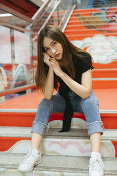 时尚写真-身穿黑色衬衫和牛仔裤裤子的泰国亚裔年轻女子坐在红色楼梯上涂鸦。
