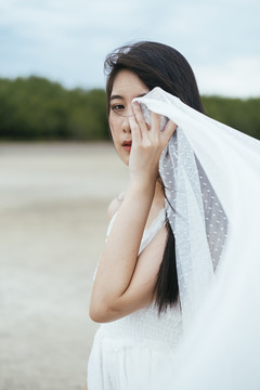 特写-年轻的亚洲长黑发女子新娘的肖像，戴着白色面纱，在户外用手遮住眼睛。
