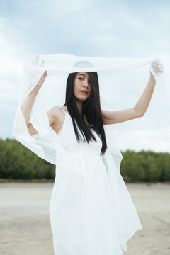 年轻的亚洲长发黑发女子新娘在户外戴着白色面纱的肖像。刮风的日子。乡村背景。