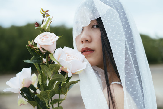 特写-年轻的亚洲长黑发女子新娘与白色面纱举行室外白玫瑰花肖像。