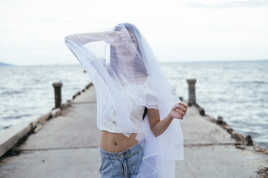 年轻的亚洲长发黑发女子戴着白纱在海桥上的肖像。衬衫和蓝色牛仔裤长裤。