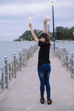 苗条的女孩。杜什时间，一位年轻的泰国亚裔女子双手捧着两个火花在海边散步。