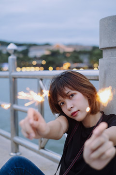 年轻的泰国亚裔女子短发坐在海桥上玩火花在黄昏时间。