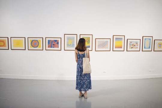 后视图-美丽的时髦的亚洲妇女在蓝色的裙子在墙上看艺术作品。美术馆。