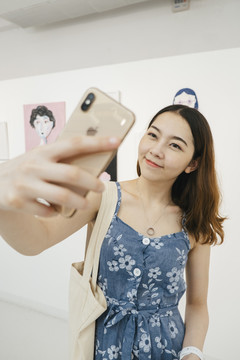 身着蓝色连衣裙的亚洲时髦美女在美术馆内与智能手机自拍。