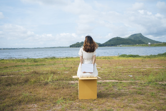 后视图-年轻的亚洲泰国妇女时髦卷发白色礼服举行空白纸的盒子后面的标志在湖。复制空间。