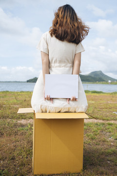 后视图-年轻的亚洲泰国妇女时髦卷发白色礼服举行空白纸的盒子后面的标志在湖。复制空间。