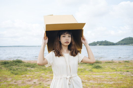 年轻的泰国亚裔女子嬉皮卷发的白色连衣裙玩纸板箱放在头上在湖边。