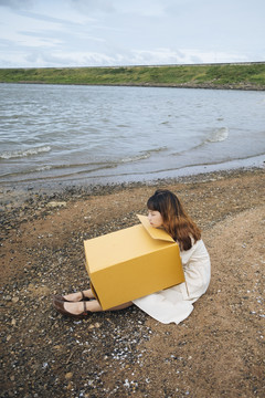 年轻的泰国亚裔女子留着时髦的卷发，穿着白色连衣裙，拿着一个纸板箱坐在湖边。