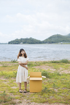 年轻的泰国亚裔女子时髦卷发，身穿白色连衣裙，手持纸板箱站在湖边。