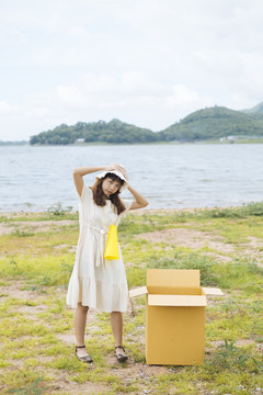 在湖边的纸板箱旁，一位年轻的泰国亚裔女子卷发，身着白色连衣裙，玩着黄色玩具扬声器扩音器。