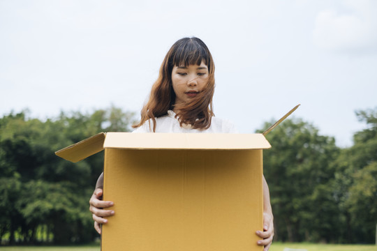 年轻的泰国亚裔女子时髦卷发，白色连衣裙，手持巨大的纸板箱。礼盒。惊讶地看着里面。