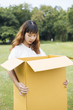 年轻的泰国亚裔女子时髦卷发，白色连衣裙，手持巨大的纸板箱。礼盒。惊讶地看着里面。