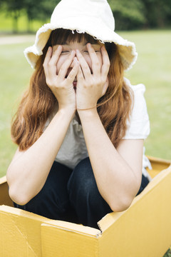 在大自然的绿色草地上，嬉皮的泰国亚裔年轻女子留着时髦的卷发，穿着白衬衫，戴着帽子，坐在纸板箱里。