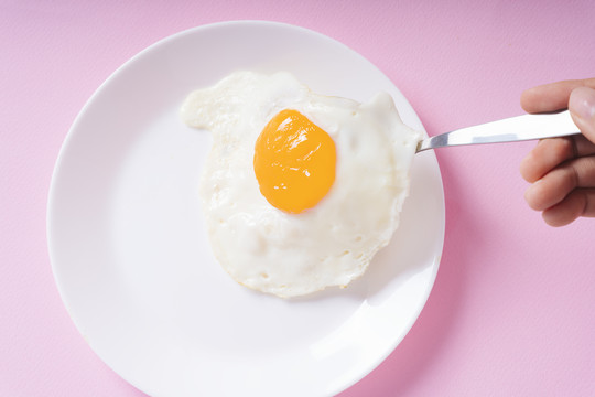 白色盘子上的煎蛋，用叉子在粉色背景上吃。