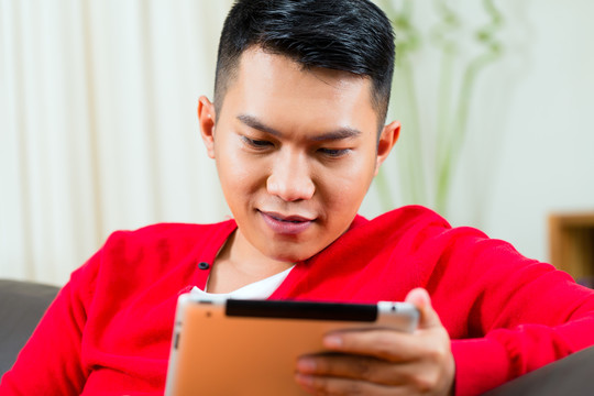 一个印尼年轻人坐在沙发上，手里拿着一台平板电脑，也许他在写东西，找电子邮件，或者在网上购物